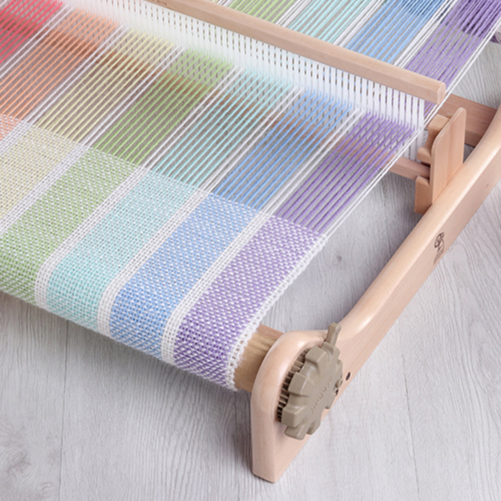Un métier Ashford à peigne envergeur en cours d’utilisation avec des fils de couleur pastel, posés sur une table de tissage